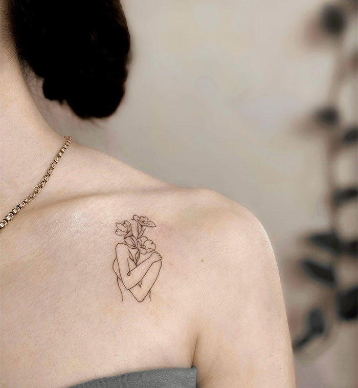 Minimalist small tattoo of a figure with flowers on a shoulder.
