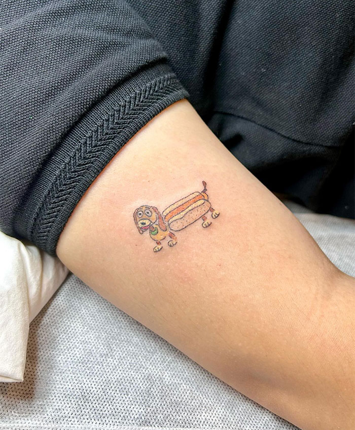 Small tattoo of a cartoon dog shaped like a hot dog on an arm.