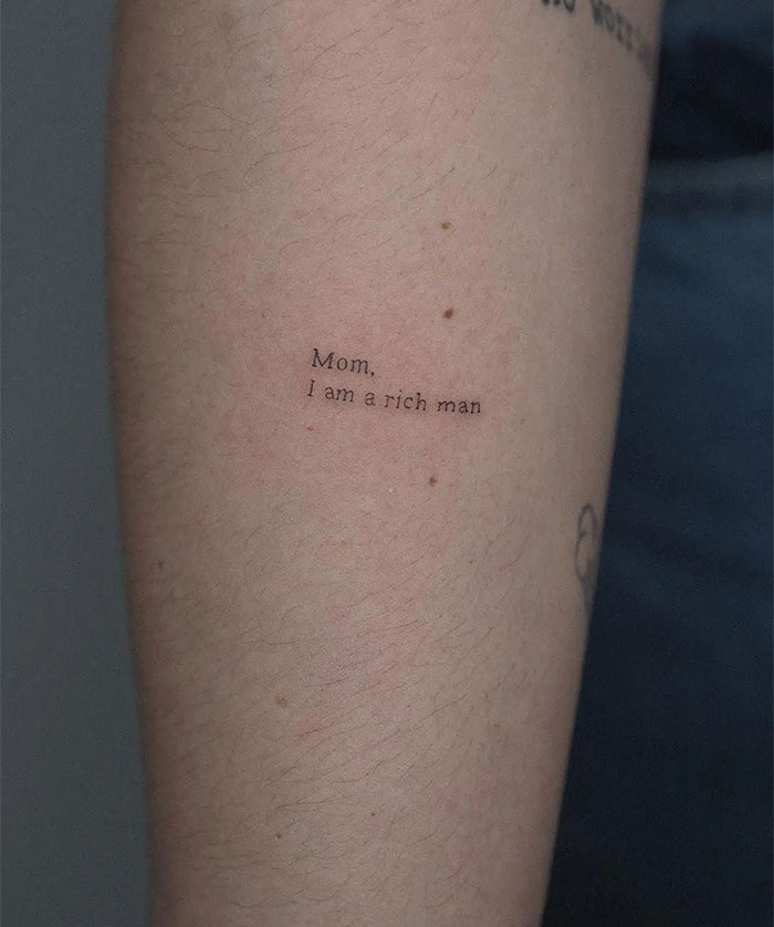 Small tattoo on an arm reading "Mom, I am a rich man" in minimalist black ink.