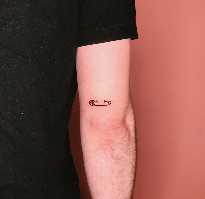 Small tattoo of a minimalist safety pin on an arm against a peach background.