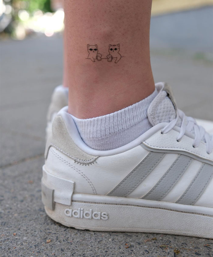Small tattoo of two cute cats on an ankle wearing white socks and sneakers.