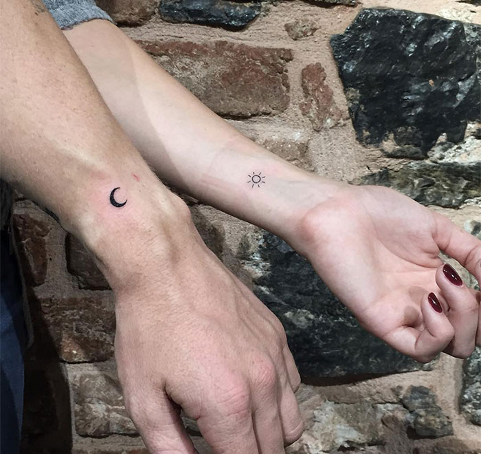 Two arms displaying small tattoos: a crescent moon and a sun, symbolizing balance, against a rustic brick background.