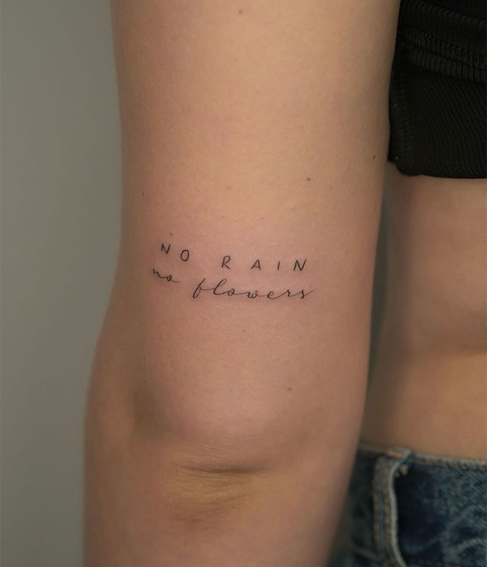 Small tattoo with the phrase "no rain no flowers" in delicate script on an upper arm.