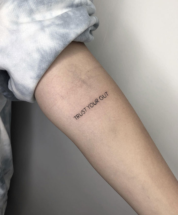 Small tattoo on arm with the words "Trust Your Gut" in minimalist font.