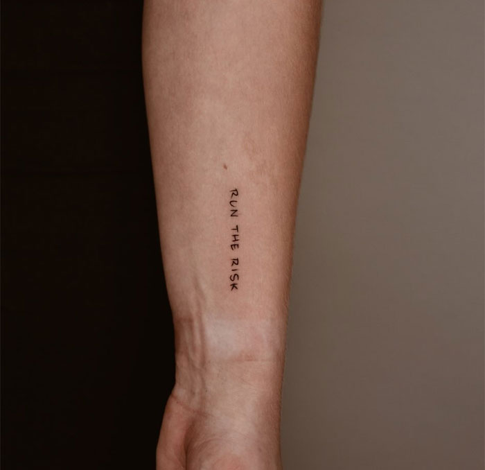 Minimalist small tattoo on forearm reading "Run the Risk," showcasing bold yet simple design.