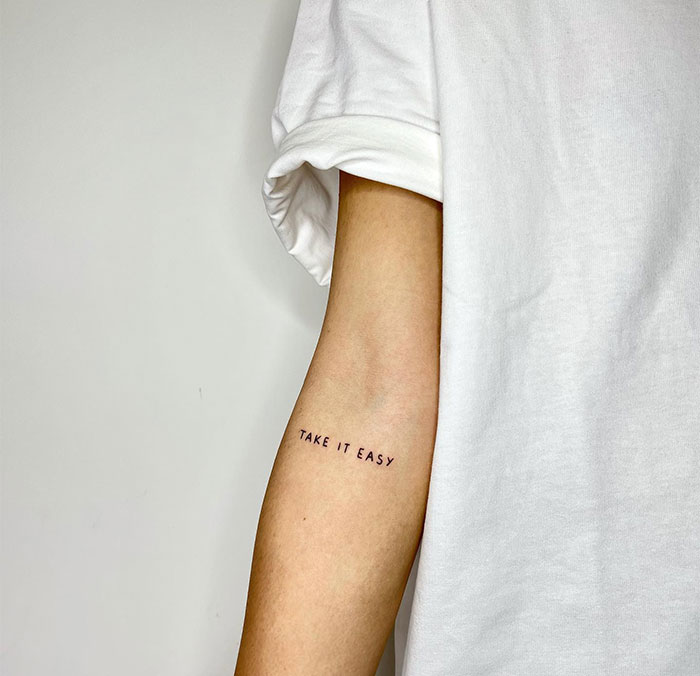 Small tattoo with "Take It Easy" text on a person's arm in a white shirt.