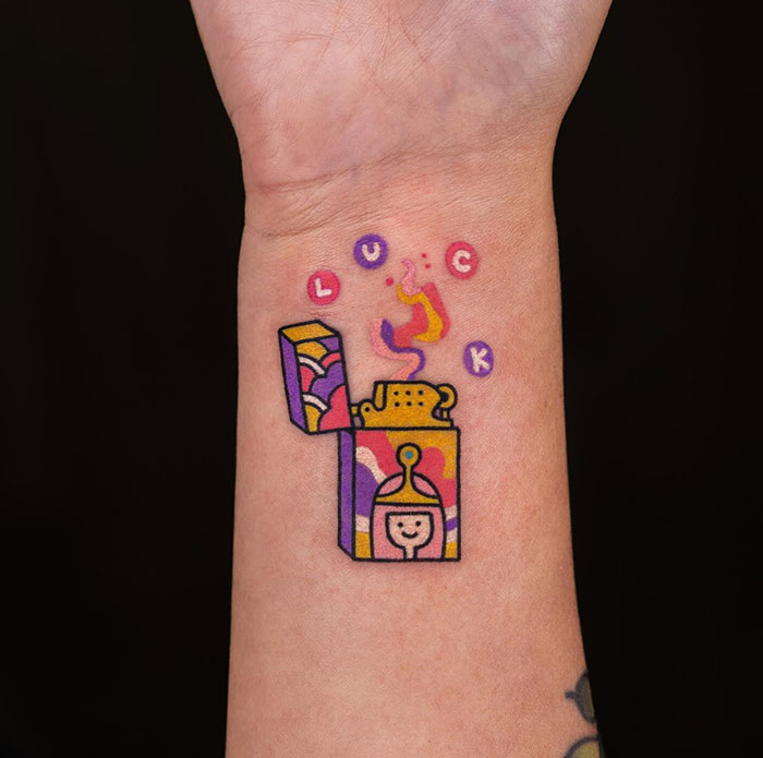 Colorful small tattoo of an open lighter with floating letters spelling "LUCK" on wrist.