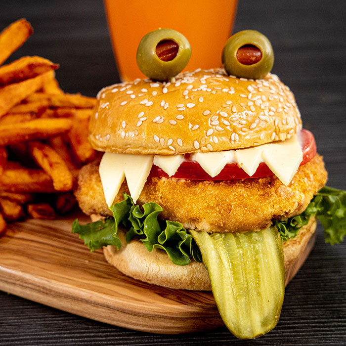 Halloween snacks featuring a monster-themed chicken sandwich with olive eyes, cheese teeth, and a pickle tongue.