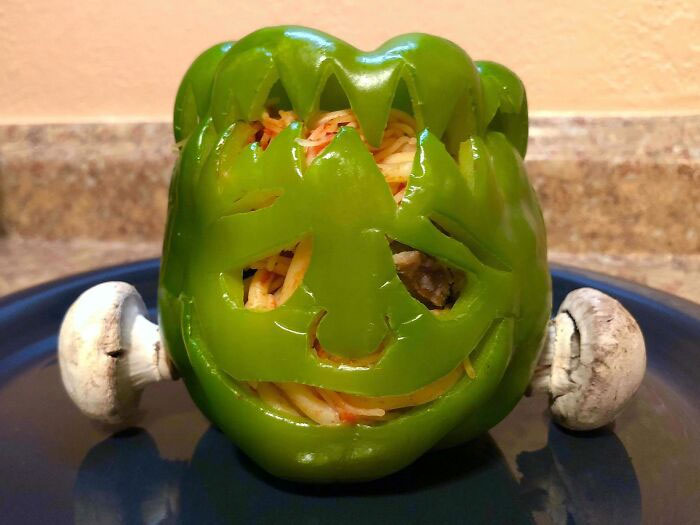 Green bell pepper carved like a jack-o'-lantern filled with spaghetti, with mushrooms on a blue plate Halloween snacks.