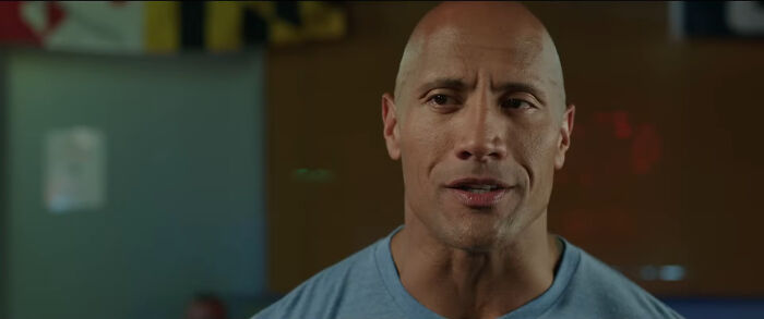 Bald man in a gray shirt, standing indoors, talking.