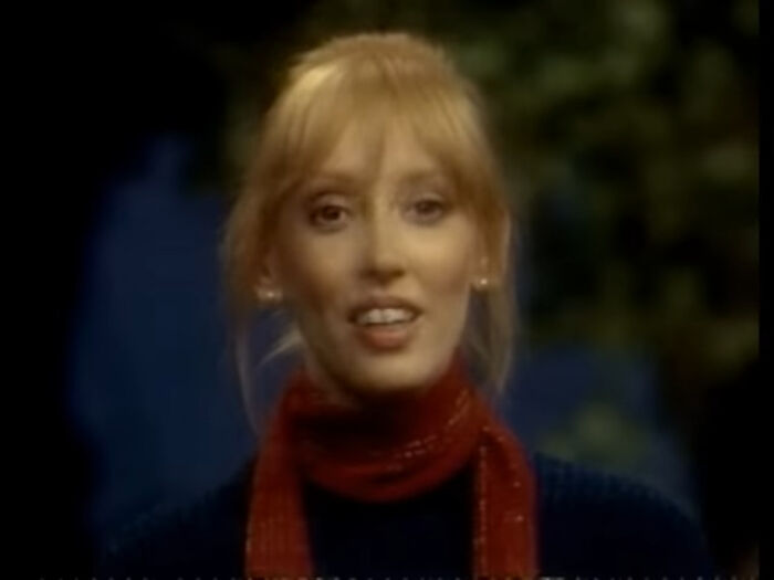 A woman with blonde hair and a red scarf smiles warmly against a blurred outdoor background, discussing actors.
