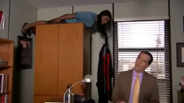 Woman performing a viral internet trend by lying horizontally on a wardrobe while a man in a suit watches.
