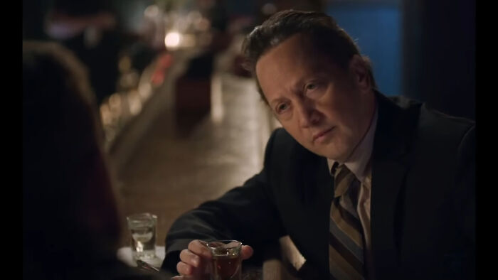 Man in a suit holding a drink at a bar, looking thoughtfully in a dimly lit atmosphere.