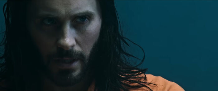 A person with long hair and a beard in an intense scene, wearing an orange shirt, related to actors in movies.