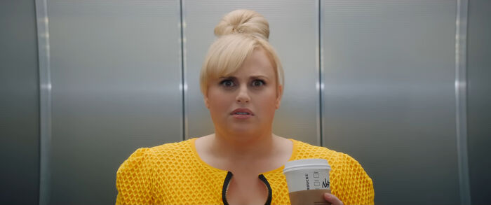 Actor in a yellow outfit holding a coffee cup in an elevator, looking surprised.