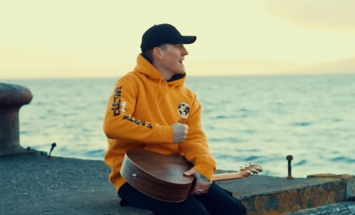 Person in an orange hoodie and black cap sitting by the ocean holding a guitar, representing trends that disappeared quickly.