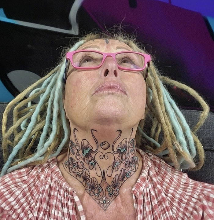 A woman with face tattoos and pink glasses, wearing a red-checkered shirt, looking upwards.