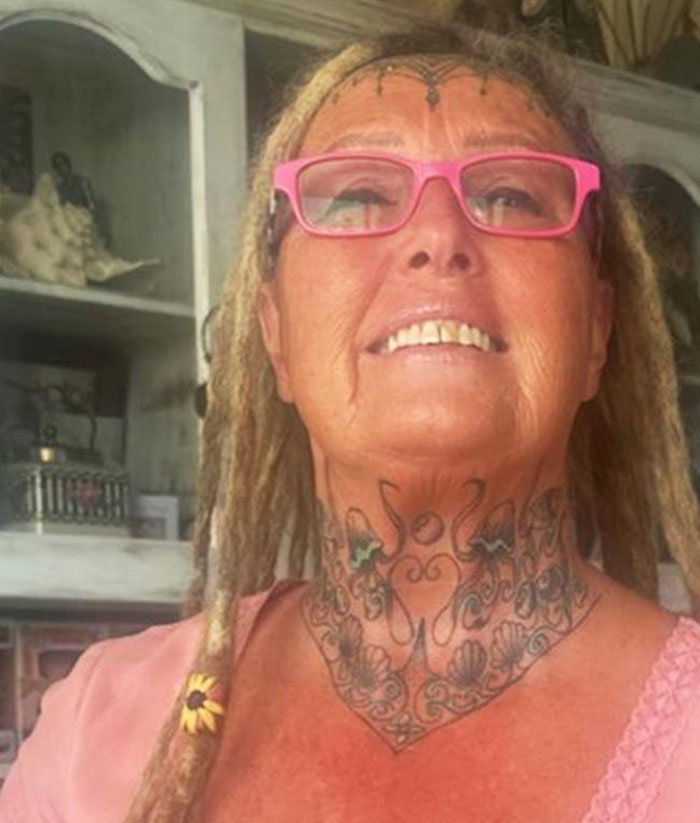 63-year-old woman with face tattoos and pink glasses smiling indoors.