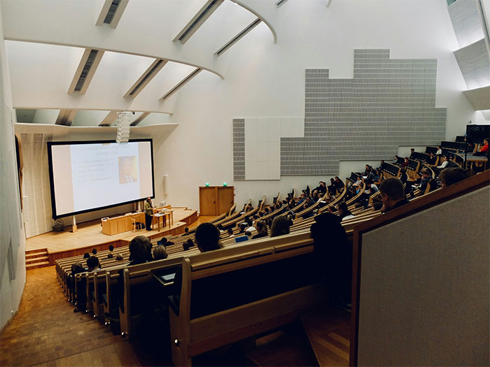 College lecture hall with students listening to a professor, focusing on college dynamics and academic challenges.