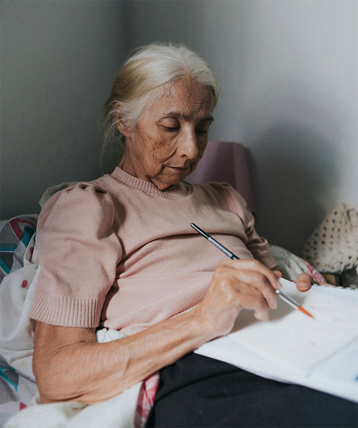 Elderly woman writing, revealing family secret in her final days, wearing a pink sweater and seated comfortably. Elderly woman writing, revealing family secret in her final days, wearing a pink sweater and seated comfortably.