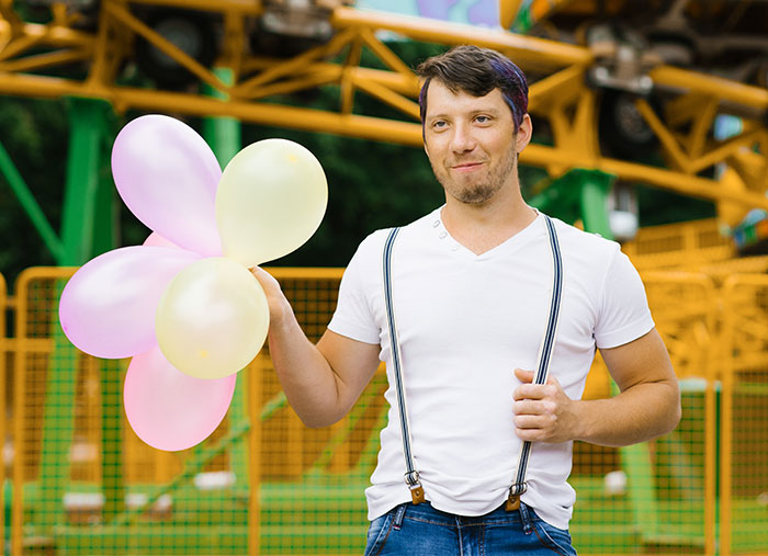 Man holding balloons at a fairground, dressed in a white shirt and suspenders, smiling about a prank. Man holding balloons at a fairground, dressed in a white shirt and suspenders, smiling about a prank.