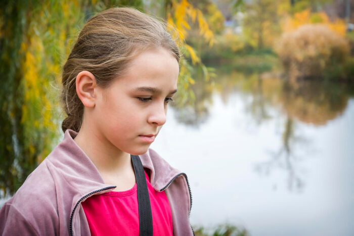 A young girl looks thoughtful by the pond, wearing a pink shirt and a brown jacket, surrounded by autumn foliage.
