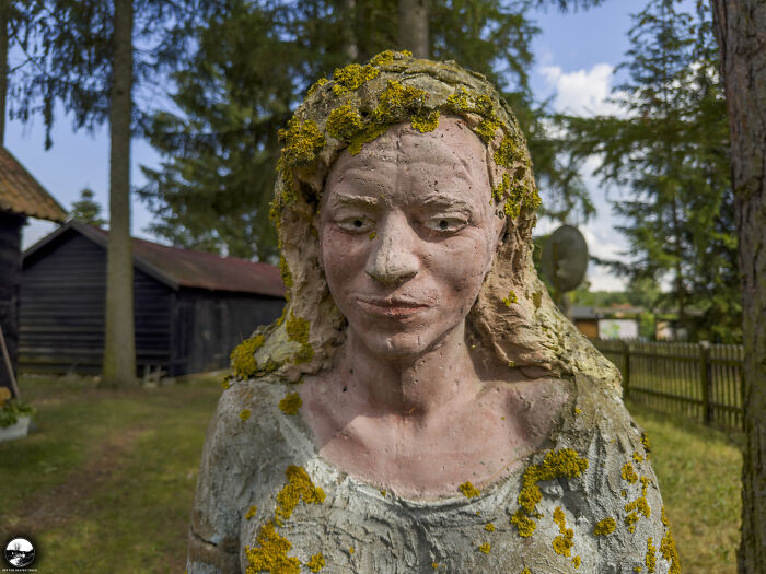 A sculpture by Adam Szubski depicting a woman with moss-covered hair in a wooded outdoor setting.