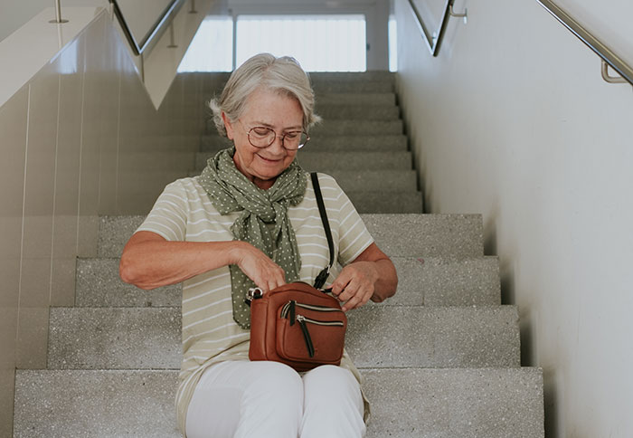 Elderly woman sitting on stairs, searching through her brown handbag, wearing glasses and a green scarf.