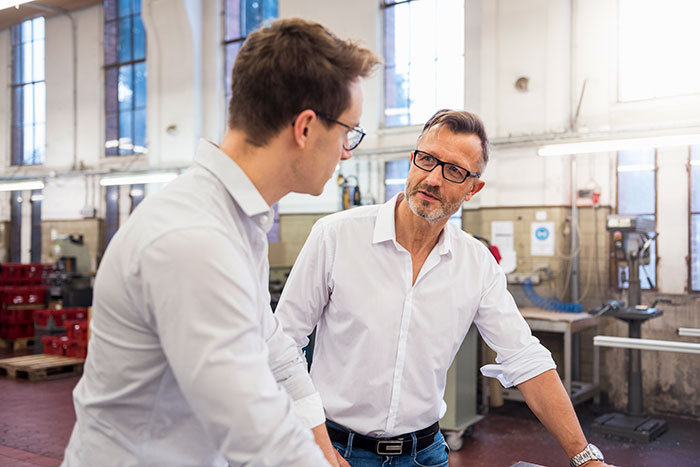 Two men in a factory setting, engaged in a discussion about input and feedback. Two men in a factory setting, engaged in a discussion about input and feedback.