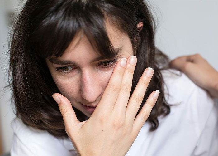 Woman in emotional distress after hearing a savage insult, holding her hand to her face in a white shirt.