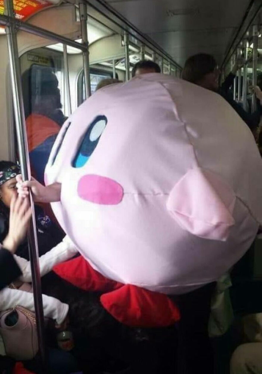 Person wearing a large, round character costume on a crowded subway, embodying "pics that go hard" in a playful setting.