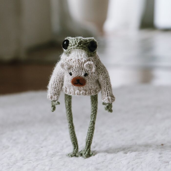 Miniature knitted frog by French artist wearing a sweater with a bear design, standing on a textured white surface.