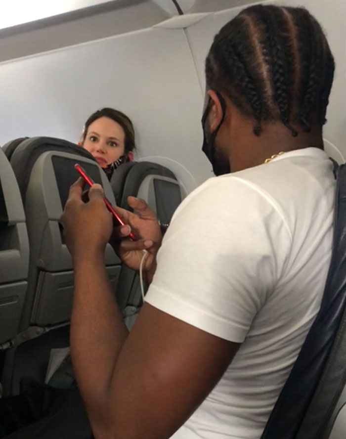 Passenger using a phone on an airplane, with another passenger looking annoyed in the background.