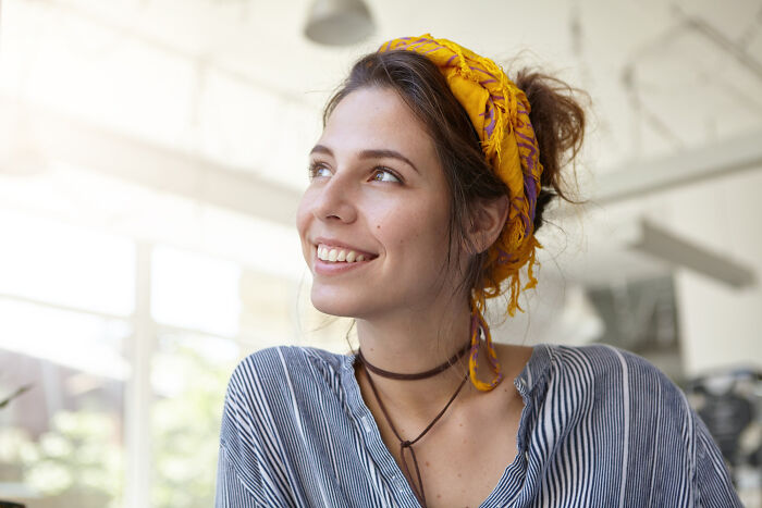 Woman smiling indoors, wearing a striped shirt and a yellow headscarf, illustrating a moment of risky decision.