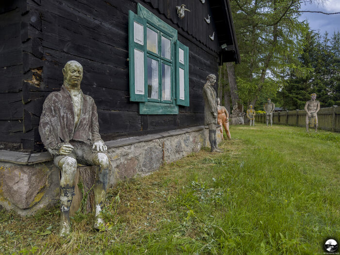 Sculptures by Adam Szubski displayed in a grassy yard, alongside a rustic wooden house with green windows.