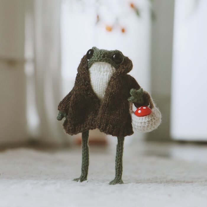 Miniature knitted frog in a coat holding a small basket indoors.