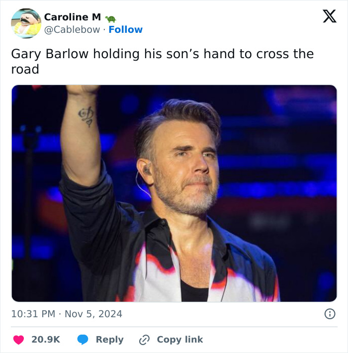 Gary Barlow on stage, raising hand, wearing a patterned shirt.