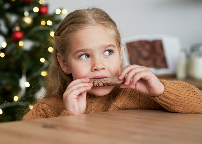 Child eating a cookie near a Christmas tree, homemade baked goods visible in the background. Child eating a cookie near a Christmas tree, homemade baked goods visible in the background.