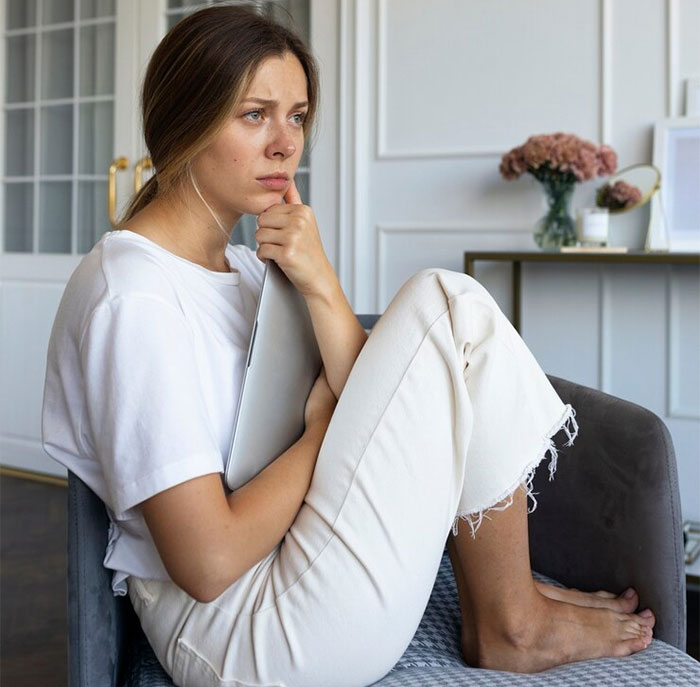 Woman sitting on a sofa, deep in thought about starting a family, holding a laptop in a cozy living room.