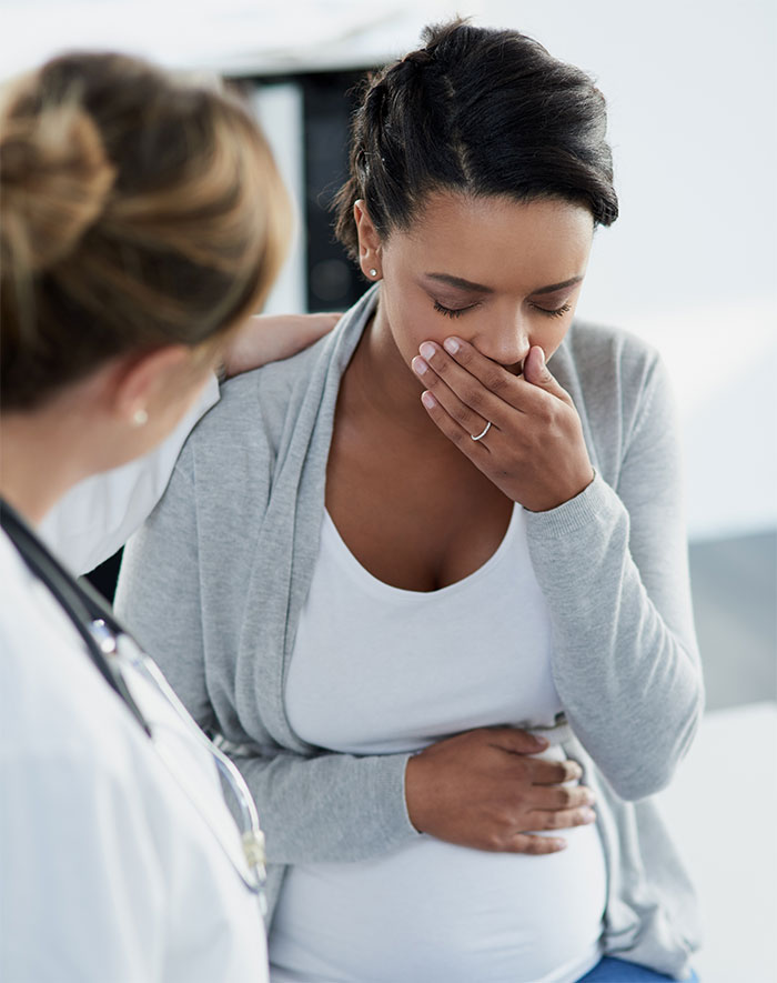 Pregnant woman in a gray cardigan holding her stomach, looking unwell, with a doctor nearby offering support.