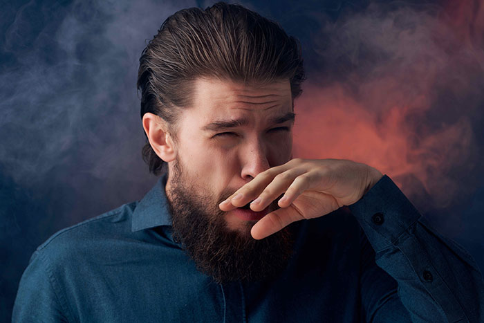 Man in a blue shirt covering his nose, surrounded by smoke, reacting to an unpleasant smell. Man in a blue shirt covering his nose, surrounded by smoke, reacting to an unpleasant smell.