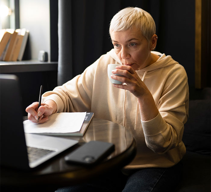 Woman in a beige hoodie sipping coffee, holding a pen, and writing notes near a laptop, conveying suspicion of MIL.