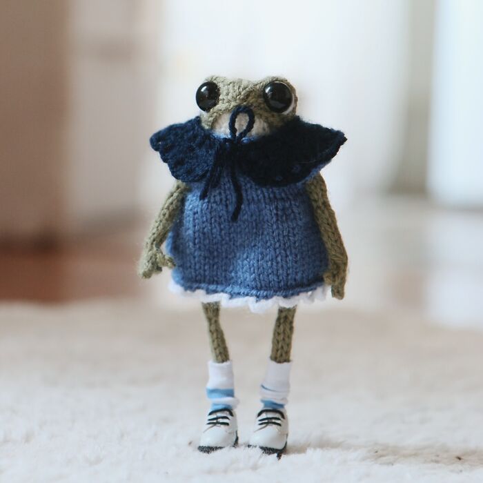 Knitted miniature frog by a French artist, wearing a blue dress and shoes, standing on a soft surface.