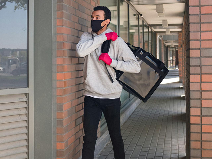 Man wearing mask and pink gloves, carrying a delivery bag near a brick building.