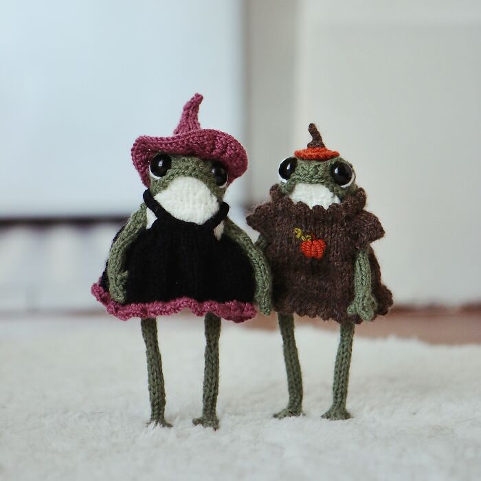 Miniature knitted frogs with hats, crafted by a French artist, standing side by side on a soft surface.