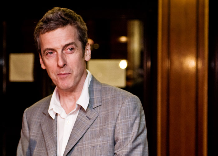 Man in a gray suit standing indoors, embodying a confident expression amidst a dimly lit background, conveying savage insult energy.