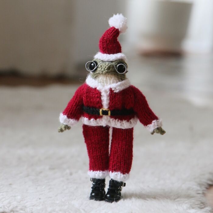 Miniature knitted frog wearing a Santa outfit with glasses, standing on a soft white surface.