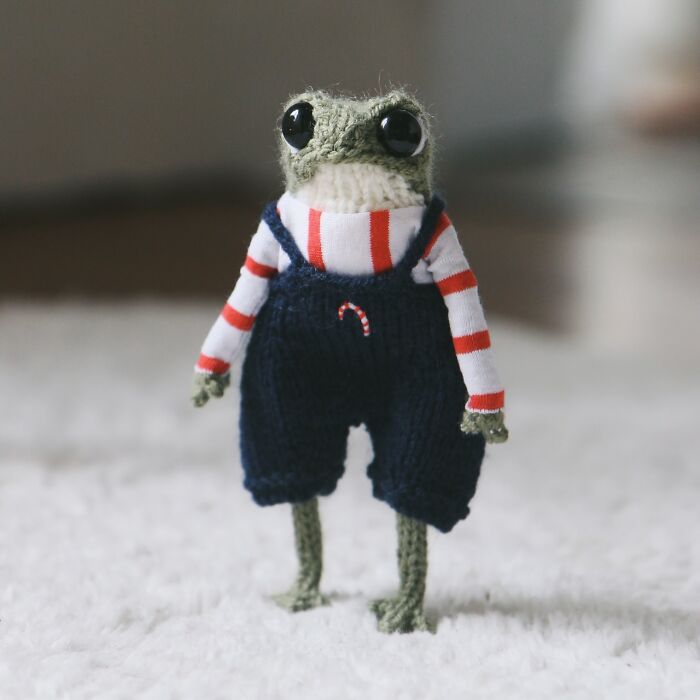 Miniature knitted frog in striped shirt and navy overalls on a soft white surface.