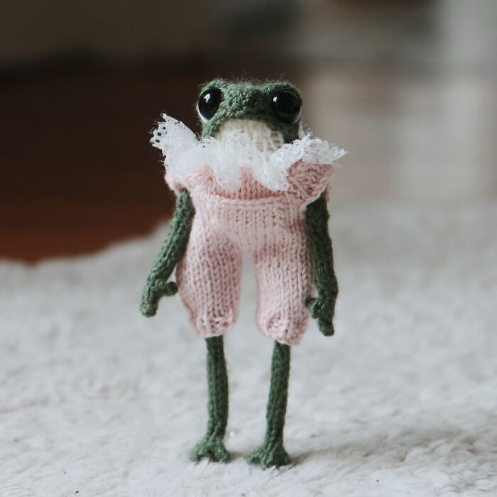 Miniature knitted frog in pink outfit with ruffled collar, created by a French artist, standing on a soft surface.