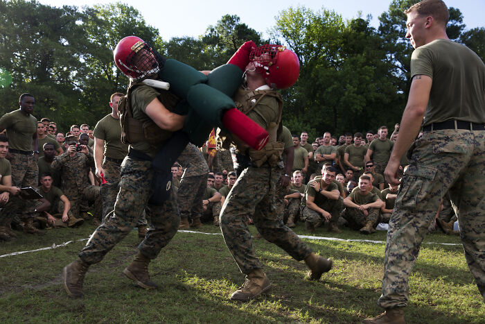 Two soldiers in combat gear spar outdoors, surrounded by fellow soldiers, demonstrating teamwork and strength.